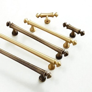 Puede incluir: Una selección de herrajes para armarios en varios acabados metálicos. La imagen muestra manijas y perillas en tonos bronce y dorado, incluyendo tiradores de barra y perillas en forma de T. El herraje está diseñado para muebles y armarios.