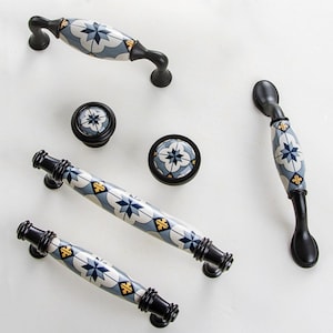 May include: A selection of cabinet handles and knobs. The black metal hardware features a ceramic centre with a blue and white geometric design and gold details. The items are displayed on a white surface. The handles and knobs are for cabinets.