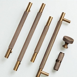 Pode incluir: Um conjunto de ferragens para armários em tom bronze, incluindo várias puxadores de barra texturizados e um botão redondo. Os puxadores variam em comprimento e a ferragem tem uma textura de padrão de diamante. Para móveis.