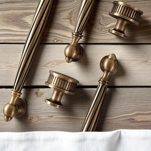 May include: A selection of antique brass cabinet handles and knobs, displayed on a light wood surface. The hardware has detailed designs with vertical grooves and rounded features, presenting a vintage aesthetic. The measurements are in centimeters.