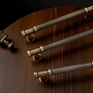 Puede incluir: Juego de herrajes para armarios en tono bronce. El conjunto incluye varios tiradores largos y acanalados y un pomo, todos con una estética vintage. El hardware se muestra sobre una superficie de madera oscura, mostrando los detalles del diseño.