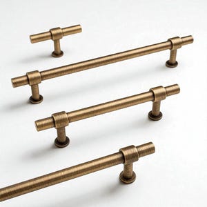 Könnte beinhalten: Eine Auswahl an bronzefarbenen Schrankgriffen und -knöpfen. Die Hardware hat ein T-Bar-Design mit einer zylindrischen Stange und einem runden Sockel. Die Oberfläche hat ein leichtes Used-Aussehen, das einen antiken oder Vintage-Stil suggeriert. Die Griffe sind unterschiedlich groß.