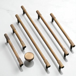 May include: A collection of bronze-colored cabinet hardware, including several bar pulls of varying lengths and a round knob. The pulls have textured ends and are arranged on a white surface. The hardware is designed for furniture.