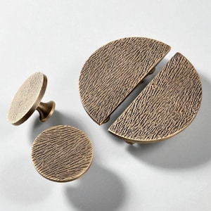 Pode incluir: Um conjunto de ferragens para armários em tom bronze, incluindo duas puxadores semicirculares, um puxador redondo e um pequeno puxador com haste. A ferragem tem uma superfície texturizada e canelada.