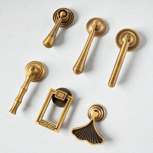 May include: A selection of antique brass cabinet handles and knobs. The collection features diverse designs, including a bamboo-style handle, a rectangular handle with a geometric pattern, and a ginkgo leaf-shaped knob. The hardware is set against a white background.
