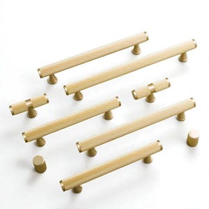May include: A collection of gold-toned cabinet hardware, including several long bar pulls and smaller knobs. The hardware features a textured, cross-hatched pattern on the main body and a smooth, rounded end. The hardware is designed for use on cabinets and drawers.