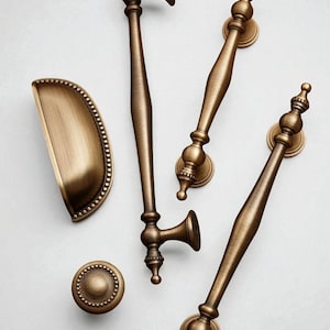 Può includere: Un assortimento di ferramenta per mobili in bronzo antico, tra cui maniglie, un pomello e una maniglia a tazza. Le maniglie variano nel design, con dettagli ornati e una finitura calda e invecchiata. La ferramenta è disposta su una superficie bianca.