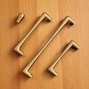 Op de afbeelding: Een set messingkleurige kastbeslagen, waaronder twee lange stanggrepen, een kortere greep en een kleine knop. De hardware is geplaatst tegen een lichte houtnerf achtergrond, met een modern design.