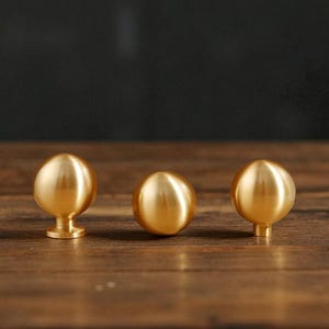 May include: Three brass cabinet knobs with a spherical design. Two knobs have a short stem and base, while the center knob is a simple sphere. The knobs are arranged on a dark wood surface.