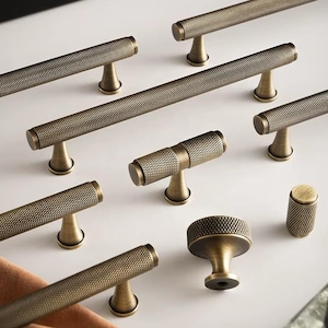 May include: Collection of antique brass cabinet hardware, including various sizes of bar pulls and a knob. The hardware features a textured, diamond-patterned surface and a warm, aged finish. The pulls are mounted on a white surface.