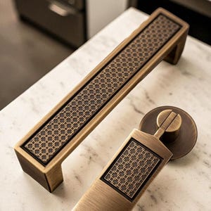 Puede incluir: Dos herrajes de gabinete en tono bronce. Uno es un mango rectangular largo con un patrón geométrico, y el otro es un tirador más pequeño con un diseño similar. Ambos tienen una estética vintage.