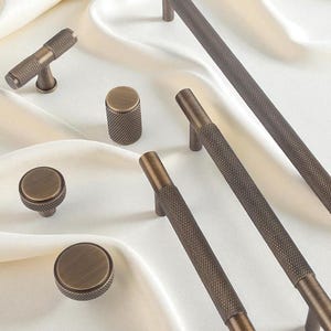 Könnte beinhalten: Eine Auswahl an bronzefarbener Schrankhardware, darunter Knöpfe und Griffe, mit einer strukturierten Oberfläche mit Rautenmuster. Die Hardware ist auf einem weißen, seidigen Stoff angeordnet und zeigt verschiedene Formen und Größen für Möbelanwendungen.