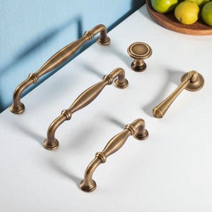 Op de afbeelding: Een assortiment antiek messing kastbeslag, waaronder verschillende handgrepen en een knop. Het beslag heeft een vintage esthetiek, met gebogen ontwerpen en een warme, metallic afwerking. De stukken zijn gerangschikt op een wit oppervlak.