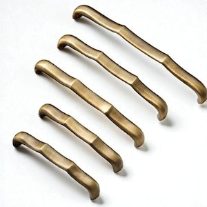 May include: Five antique brass cabinet pulls arranged diagonally. Each pull has a curved handle and a textured, bamboo-like design. The pulls are a warm, golden-brown color, and are set against a white background.