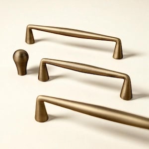 Puede incluir: Un conjunto de herrajes para armarios de color bronce, que incluye tres tiradores de barra y un pomo. Los tiradores tienen un diseño suavemente curvado con postes angulados. El pomo es esférico. Todas las piezas tienen un acabado texturizado.