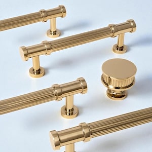 May include: A selection of gold-coloured cabinet hardware, including bar pulls and a round knob. The pulls feature a ribbed design and are mounted on small, circular bases. The knob has a similar ribbed texture and a flat top.