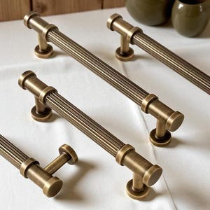 May include: Bronze-toned cabinet pulls with a ribbed design. The pulls have a cylindrical center and rounded ends, with a curved base for mounting. The hardware is arranged on a white surface, with two decorative vases in the background.