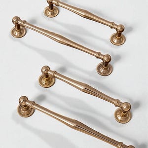 Pode incluir: Quatro puxadores dourados para armários com um design clássico. Cada puxador apresenta um centro arredondado e uma base circular decorativa. Os puxadores estão dispostos sobre um fundo branco, mostrando sua estética elegante e vintage.