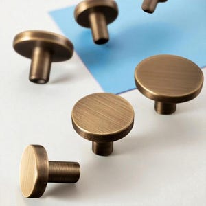 May include: Several bronze-colored cabinet knobs with a brushed finish are arranged on a white and blue surface. The knobs have a round, flat top and a cylindrical stem. The hardware is designed for furniture and home decor.