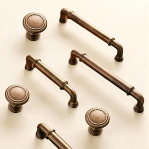 Może przedstawiać: Zestaw okuć do szafek w kolorze brązu, obejmujący okrągłe gałki i uchwyty prętowe. Gałki mają podniesiony, okrągły wzór, a uchwyty są proste z zakrzywionymi końcami. Okucia są ułożone na jasnej powierzchni.