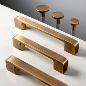 Puede incluir: Un conjunto de herrajes para gabinetes de color bronce, que incluye tres tiradores de barra rectangulares y tres pomos redondos. Los tiradores tienen un diseño simple y moderno con una forma ligeramente curvada. Los pomos son planos y circulares.