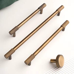 Könnte beinhalten: Bronze-farbenes Schrankbeschlagsset. Das Set enthält drei lange Stangengriffe und einen runden Knopf. Die Griffe haben an jedem Ende einen strukturierten Griff mit Rautenmuster. Hardware für Schränke und Schubladen.