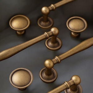 May include: Close-up of antique brass cabinet hardware, including knobs and pulls. The knobs have a round, ridged design, while the pulls have a curved handle. The hardware is set against a dark grey background.
