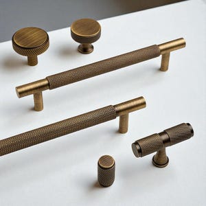 Può includere: Un assortimento di ferramenta per mobili in ottone antico, tra cui pomelli e maniglie a barra. I pomelli hanno un design rotondo e strutturato, mentre le maniglie presentano un motivo a diamante. La ferramenta è esposta su una superficie bianca.