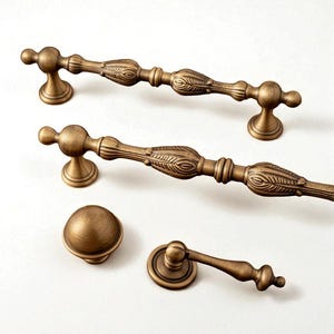 Puede incluir: Un surtido de herrajes de latón antiguo para gabinetes, que incluye dos tiradores, un pomo y un tirador pequeño. Los herrajes presentan detalles ornamentales de hojas y flores, y un acabado cálido y envejecido. Las piezas están dispuestas sobre una superficie blanca.