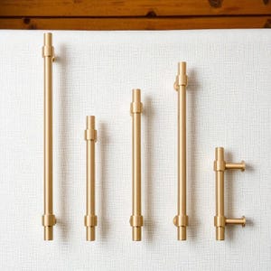 Set of modern brushed gold cabinet pull handles in multiple sizes displayed on white background.