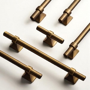 May include: A collection of bronze-colored cabinet pulls and knobs. The pulls are bar-shaped with a cylindrical center and square base. The knobs have a T-shape with a square base. The hardware is arranged on a white surface.