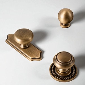 以下が含まれることがあります： 3つの異なるスタイルのアンティーク真鍮キャビネットノブ。1つはシンプルな丸いノブ、もう1つは長方形のバックプレート上の丸いノブ、3つ目はベースの周りにロープのようなデザインがあります。すべて暖かく、古びた仕上がりです。