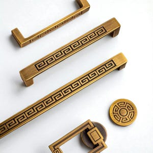 May include: Bronze-coloured cabinet hardware featuring a Greek key design. The image displays various handles and a circular knob, all with a matching decorative pattern. The hardware is likely metal and has a vintage or antique appearance.