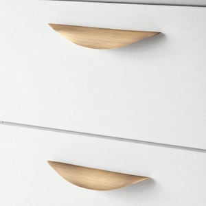 Op de afbeelding: Close-up van twee witte kastladen met gebogen, goudkleurige metalen handgrepen. De handgrepen zijn horizontaal georiënteerd en hebben een glad, modern design. De laden zijn schoon en minimalistisch.