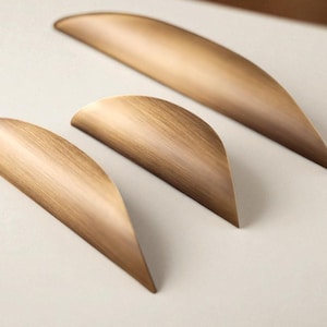 May include: Three bronze cabinet pulls on a light background. The pulls have a curved, leaf-like shape with a brushed metal texture. The hardware is designed for furniture and home decor.