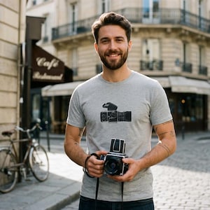 May include: A heather gray short-sleeve t-shirt featuring a black and white graphic illustration of a medium format camera on the chest. The shirt has a crew neckline and a standard fit, showcasing a detailed print of a vintage-style camera.