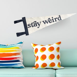 Könnte beinhalten: Hellblaues Sofa mit drei dekorativen Kissen und einem Wimpel mit der Aufschrift "stay weird". Die Kissen haben farbenfrohe Designs, darunter Streifen, Tupfen und Farbspritzer. Ein Regenbogen-Teppich liegt auf dem Boden.