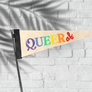 Puede incluir: Un banderín blanco con la palabra "QUEER" en letras arcoíris y la palabra "af" en letras rojas. El banderín está colgado en una pared de ladrillo blanco.