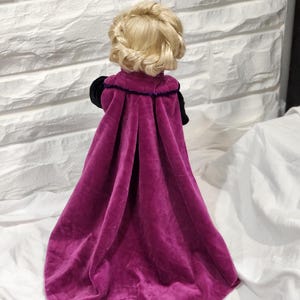 May include: A doll with blonde hair styled in a braided crown, wearing a long, magenta velvet cape with black sleeves and a purple trim. The doll is positioned with its back to the viewer, showcasing the cape's flowing design.