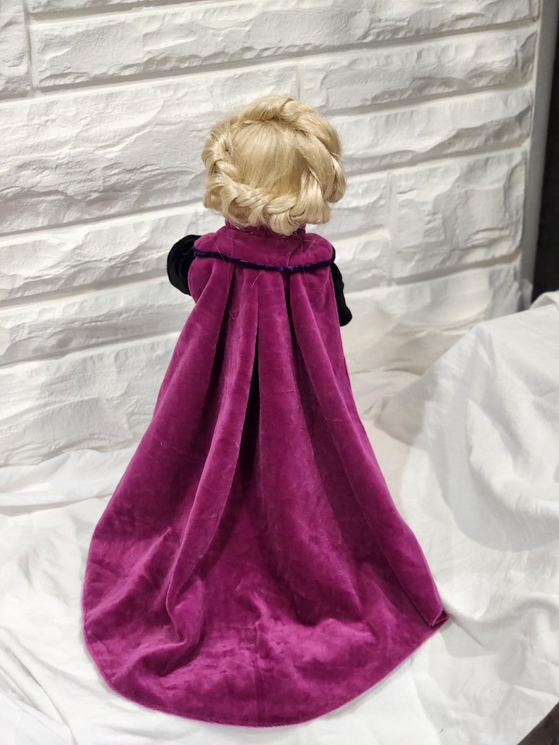 May include: A doll with blonde hair styled in a braided crown, wearing a long, magenta velvet cape with black sleeves and a purple trim. The doll is positioned with its back to the viewer, showcasing the cape's flowing design.