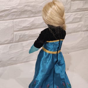 Peut inclure: Poup&eacute;e blonde coiff&eacute;e en chignon bas, v&ecirc;tue d'une robe noire et turquoise avec des bordures dor&eacute;es. La jupe turquoise est orn&eacute;e de broderies florales. Les gants de la poup&eacute;e sont turquoise.