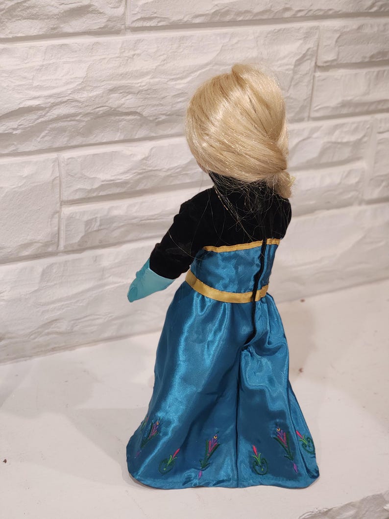Peut inclure: Poup&eacute;e blonde coiff&eacute;e en chignon bas, v&ecirc;tue d'une robe noire et turquoise avec des bordures dor&eacute;es. La jupe turquoise est orn&eacute;e de broderies florales. Les gants de la poup&eacute;e sont turquoise.