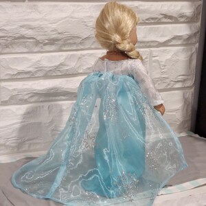 K&ouml;nnte beinhalten: Ein blaues und wei&szlig;es Puppenkleid mit einem glitzernden Umhang und Schneeflockenmuster. Das Kleid ist aus durchsichtigem Stoff und hat einen langen Schleier.