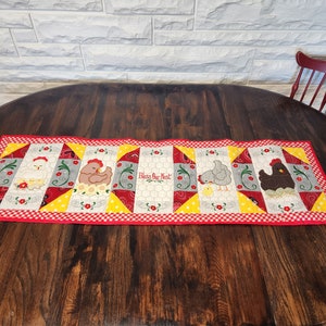 May include: A rectangular table runner with a red gingham border. The center features embroidered chickens and the text "Bless Our Nest". The design includes red, yellow, gray, and white colors, with floral accents.
