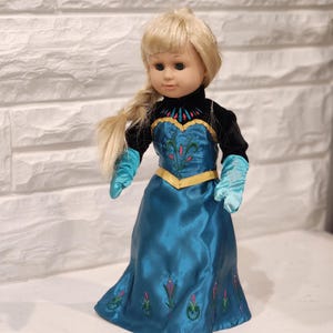 Peut inclure: Poup&eacute;e blonde avec une tresse lat&eacute;rale, v&ecirc;tue d'une robe de satin bleu sarcelle avec des d&eacute;tails floraux brod&eacute;s. La poup&eacute;e a un haut en velours noir, des gants bleu sarcelle et une bordure dor&eacute;e. La poup&eacute;e est devant un mur de briques blanches.