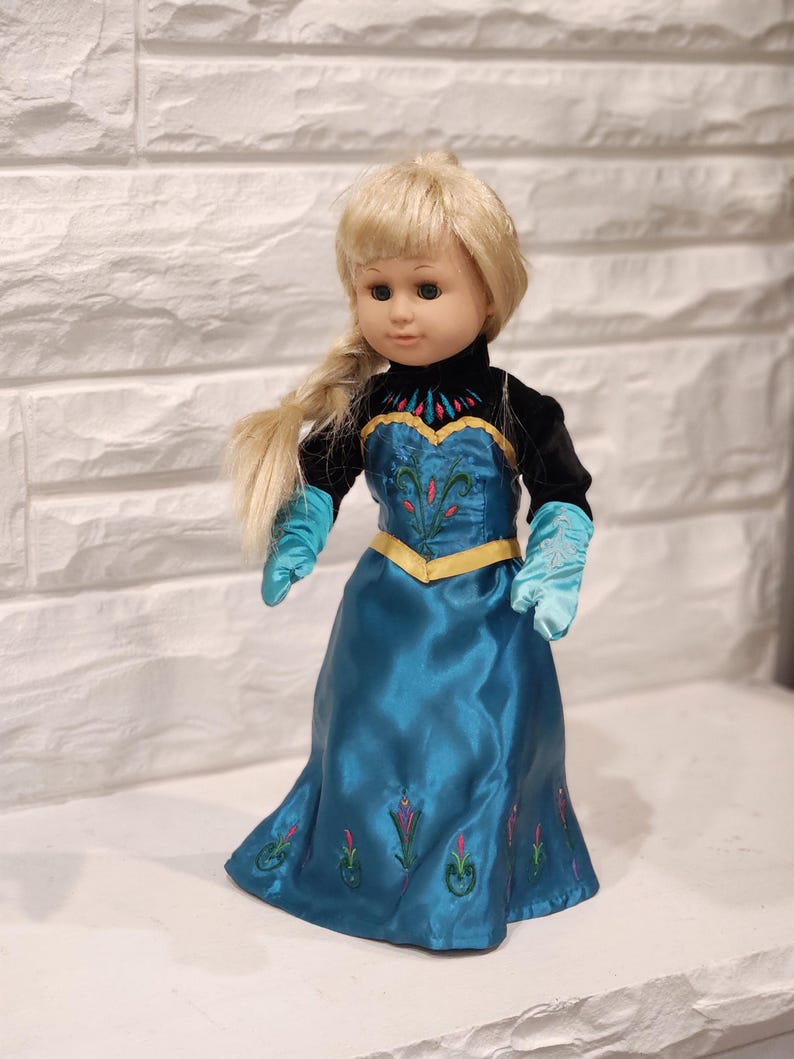 Peut inclure: Poup&eacute;e blonde avec une tresse lat&eacute;rale, v&ecirc;tue d'une robe de satin bleu sarcelle avec des d&eacute;tails floraux brod&eacute;s. La poup&eacute;e a un haut en velours noir, des gants bleu sarcelle et une bordure dor&eacute;e. La poup&eacute;e est devant un mur de briques blanches.