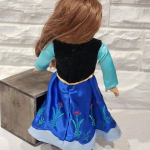 Pu&ograve; includere: Bambola con un vestito di raso blu con ricami floreali, un gilet nero e una maglia azzurra a maniche lunghe. La bambola ha lunghi capelli rossi. La bambola &egrave; accanto a una scatola di legno.