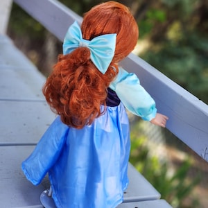 May include: A doll with long, curly red hair styled in a half-up ponytail secured with a light blue satin bow. The doll wears a dress featuring a dark purple bodice and long, light blue sleeves, paired with a flowing, solid light blue skirt.