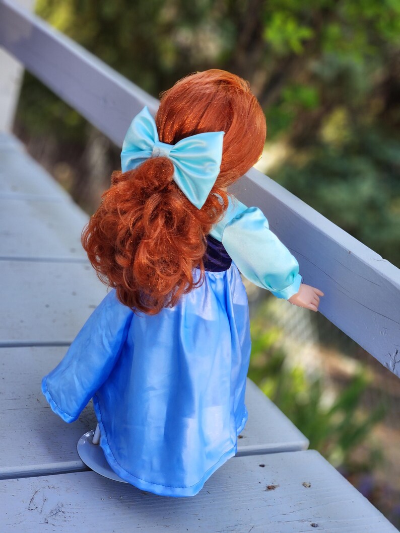 May include: A doll with long, curly red hair styled in a half-up ponytail secured with a light blue satin bow. The doll wears a dress featuring a dark purple bodice and long, light blue sleeves, paired with a flowing, solid light blue skirt.