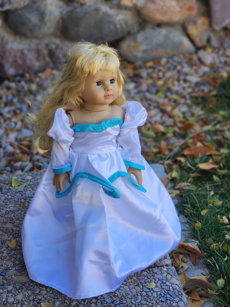 May include: A blonde doll wearing a white dress with a blue sash and a blue bodice. The doll is standing on a stone surface with leaves and grass in the background.
